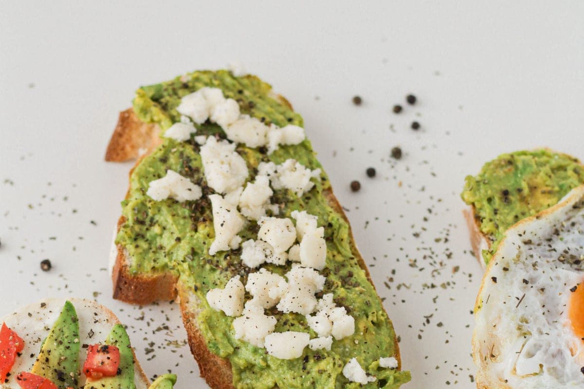 Slices of sourdough avocado toast topped with crumbled feta and cracked black pepper, with tomato-topped slices alongside