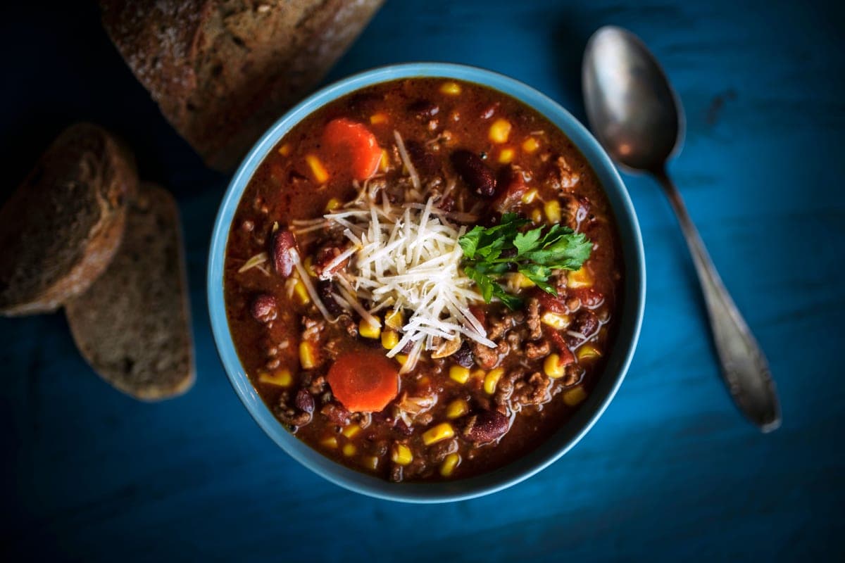 A bowl of thick homemade beef chili topped with shredded cheese and fresh herbs, with a spoon and slices of bread alongside