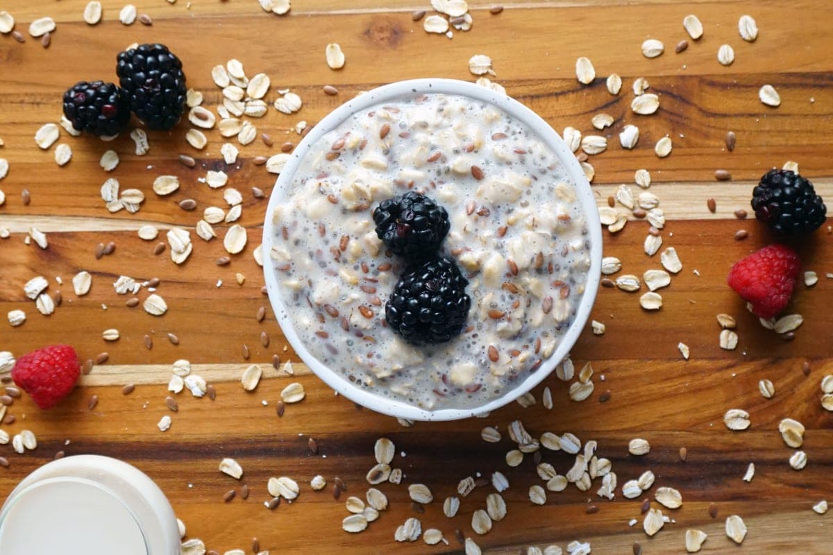A bowl of creamy overnight oats topped with fresh blackberries and raspberries on a wooden table scattered with rolled oats