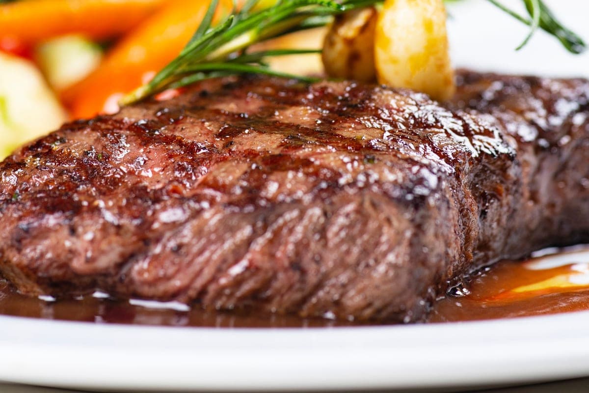 A sliced pan-seared steak with a dark crust and rosy pink center, topped with fresh rosemary and served with pan sauce and roasted vegetables