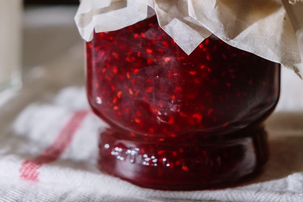 A glass jar of deep red homemade raspberry jam with a paper cover on a striped kitchen towel