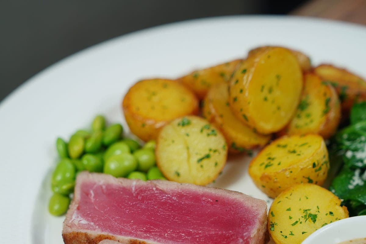 Sliced seared ahi tuna steak with a golden crust and rare pink center on a white plate with roasted potatoes and edamame