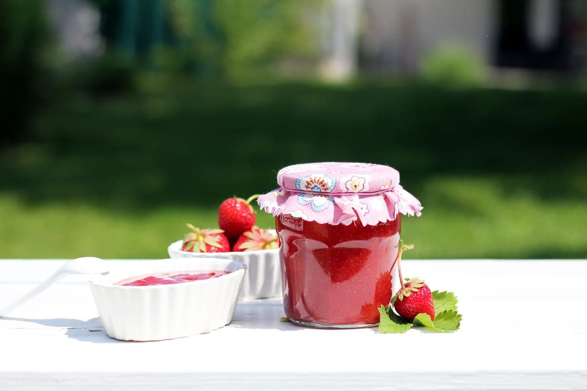 Glass jars of bright red homemade strawberry jam with fresh strawberries on a white table outdoors