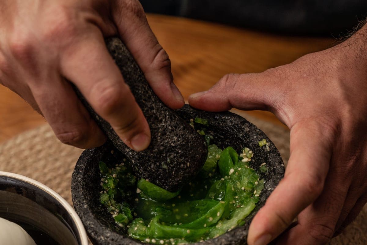 Hands grinding fresh green tomatillo salsa with a stone pestle in a traditional molcajete