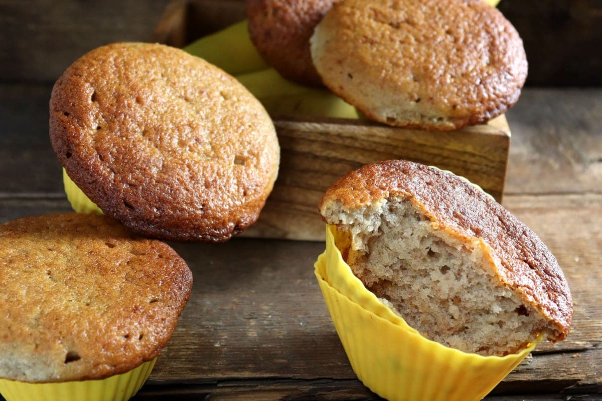 Golden banana muffins in yellow liners on a rustic wooden table, one broken open to show the soft crumb, with bananas behind