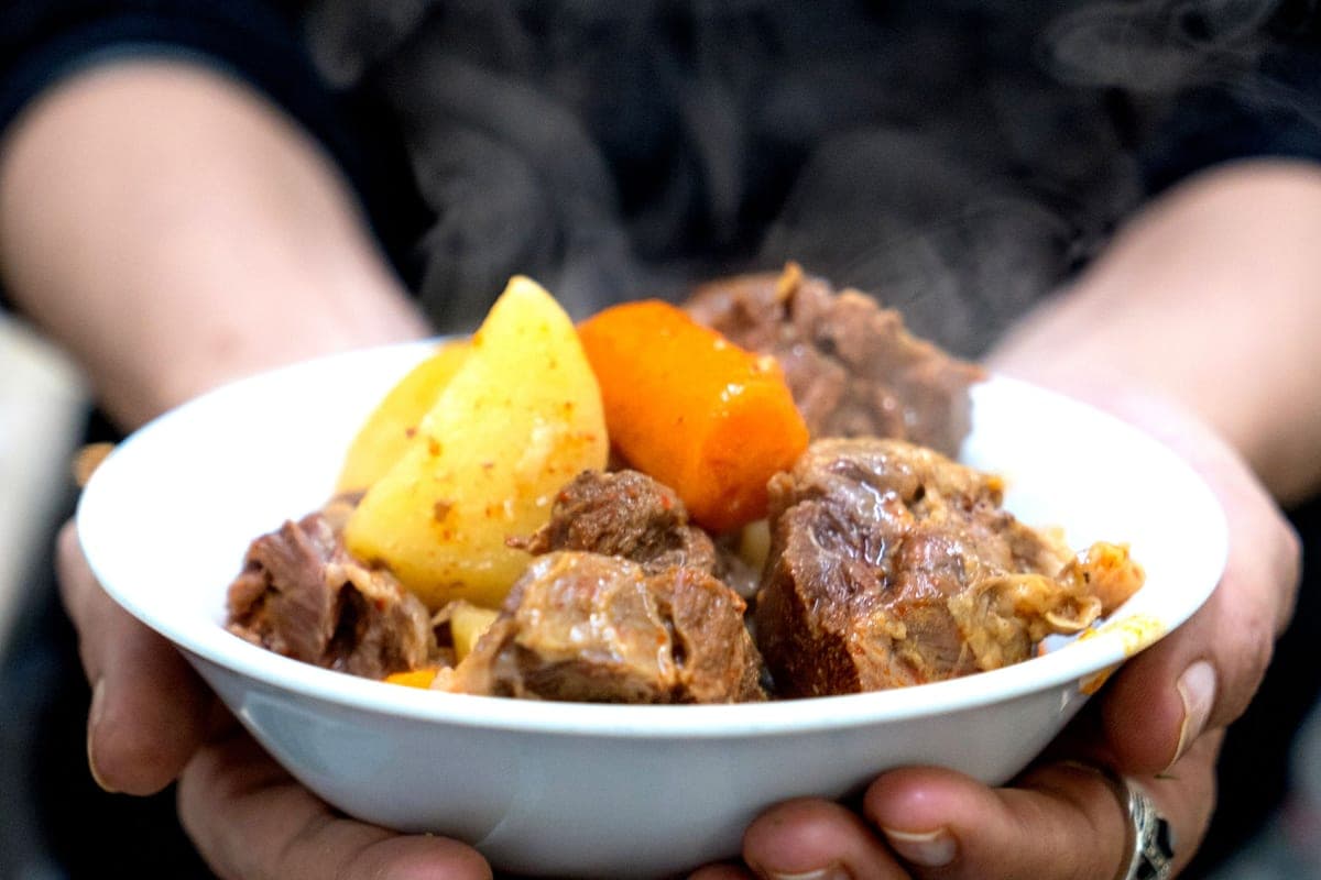 Two hands holding a steaming white bowl of beef stew with chunks of beef, potato and carrot