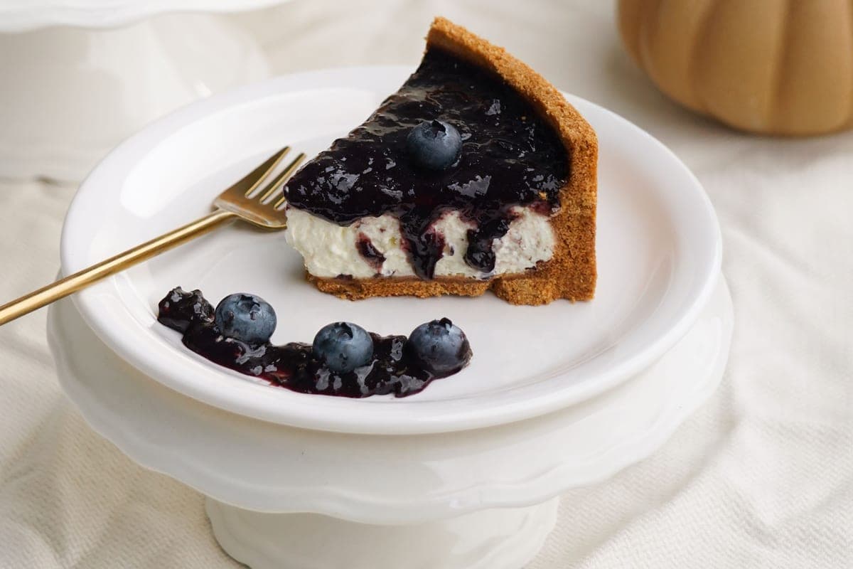 A slice of creamy baked blueberry cheesecake with a graham cracker crust and glossy blueberry topping on a white plate with fresh blueberries