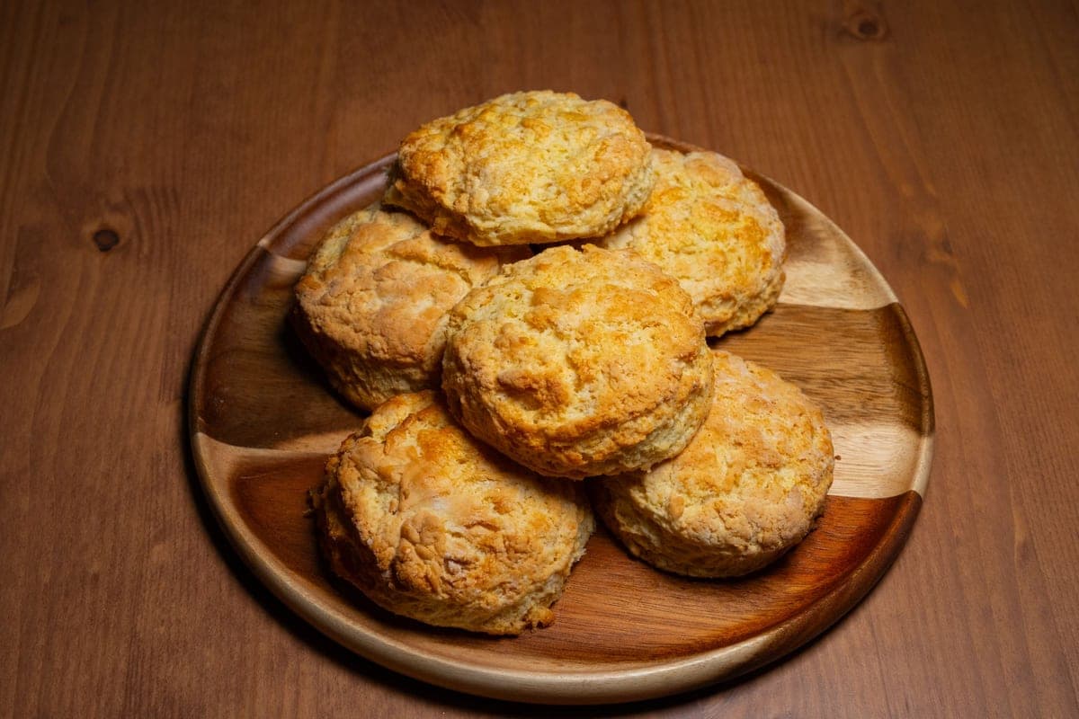 A wooden plate piled with golden, flaky buttermilk biscuits on a wooden table