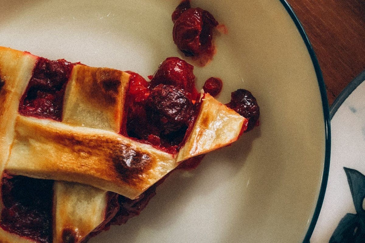 A slice of cherry pie with a golden lattice crust and whole red cherries on a cream-colored plate