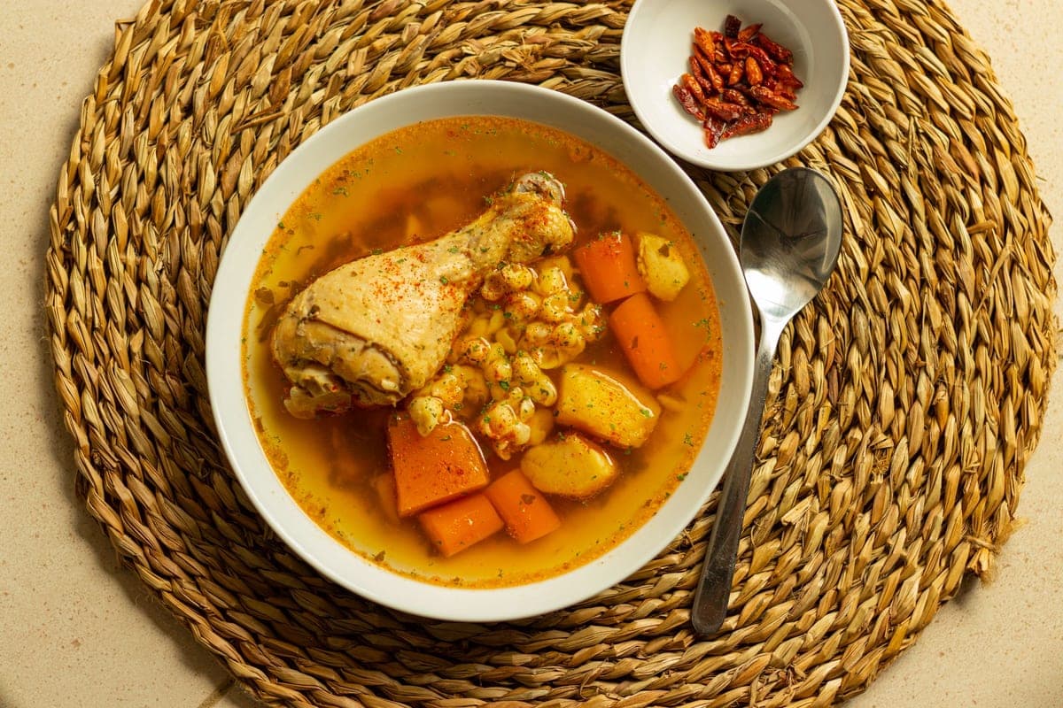 A bowl of golden chicken soup with a chicken drumstick, carrots, potatoes and corn, set on a woven placemat with a spoon