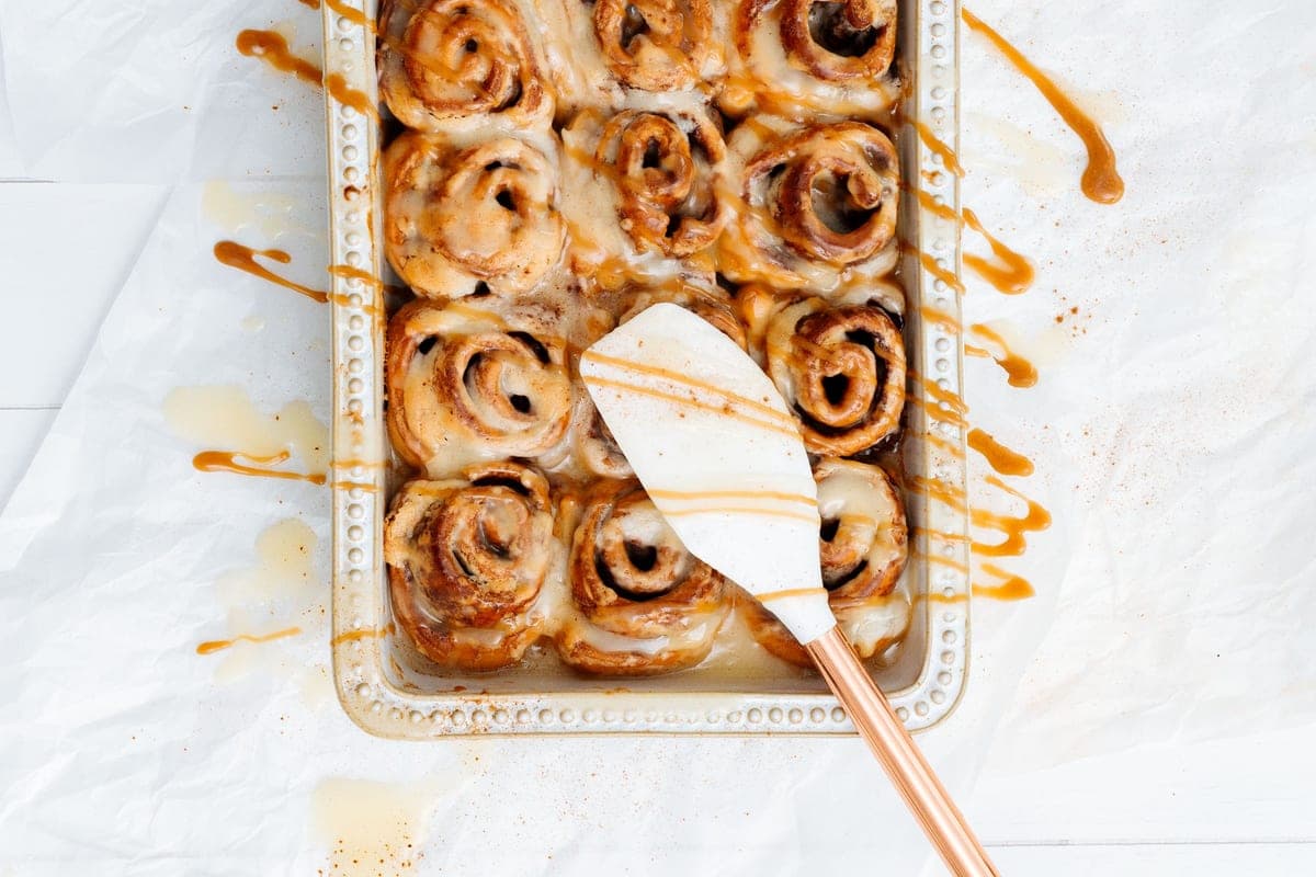 A baking dish of golden cinnamon rolls drizzled with caramel icing, with a spatula lifting one out, on white parchment