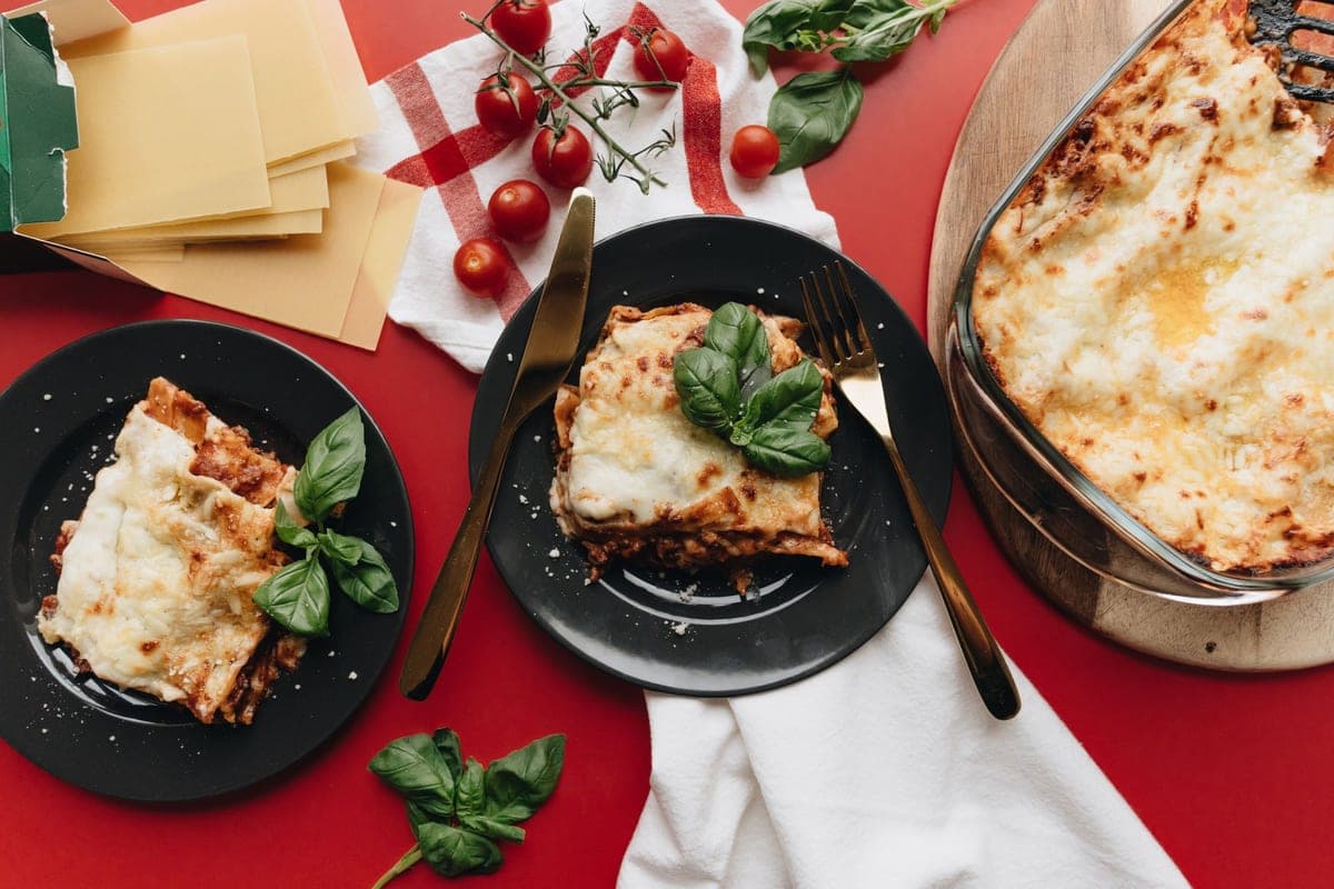 Two plated squares of lasagna garnished with basil beside the baking dish, on a red tablecloth with fresh tomatoes and dried lasagna sheets