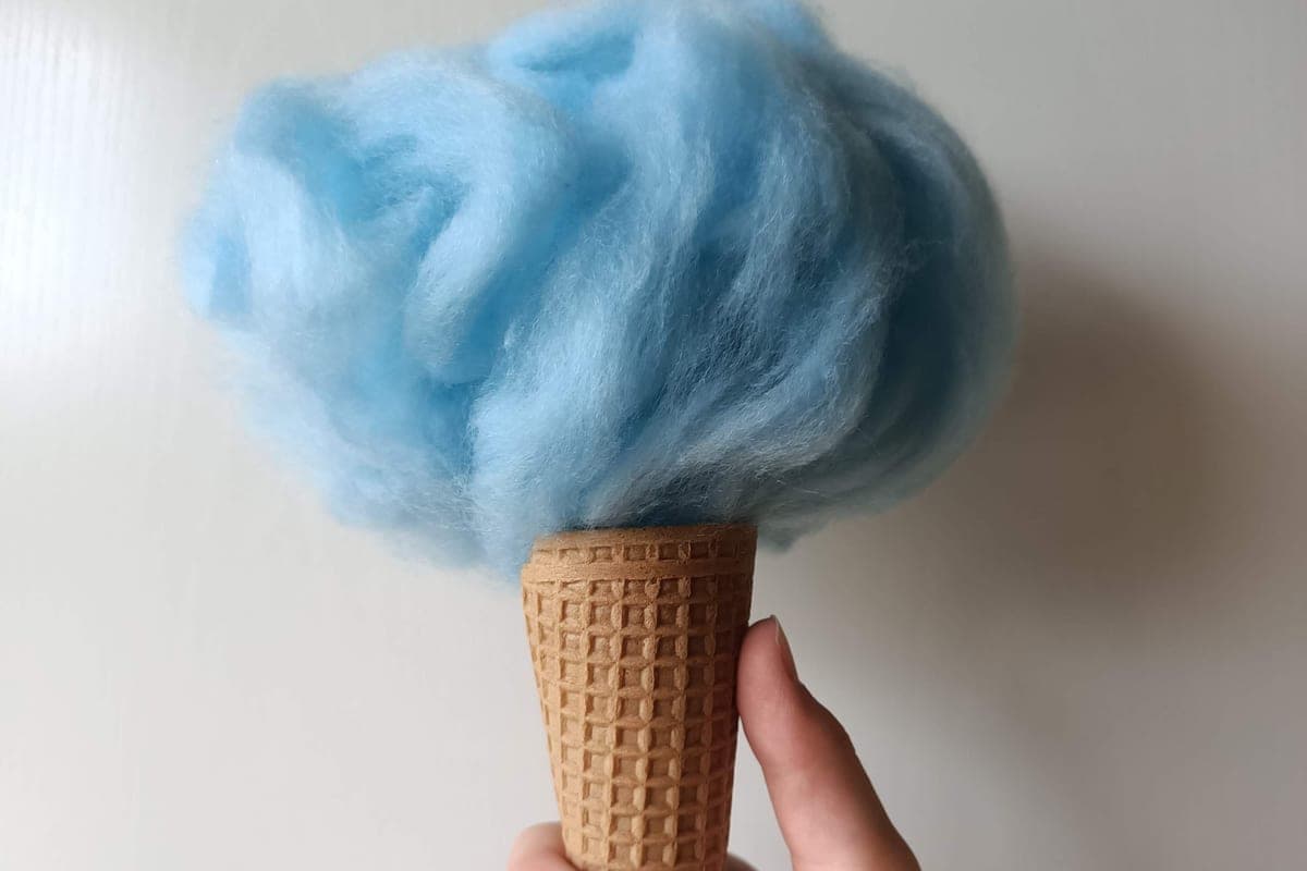 A hand holding a waffle cone topped with a fluffy cloud of blue cotton candy