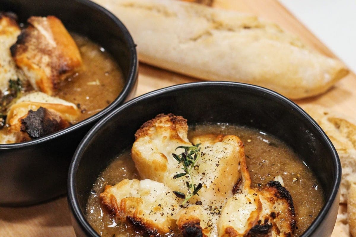 Two black bowls of French onion soup topped with toasted bread and melted cheese, with a baguette on a wooden board