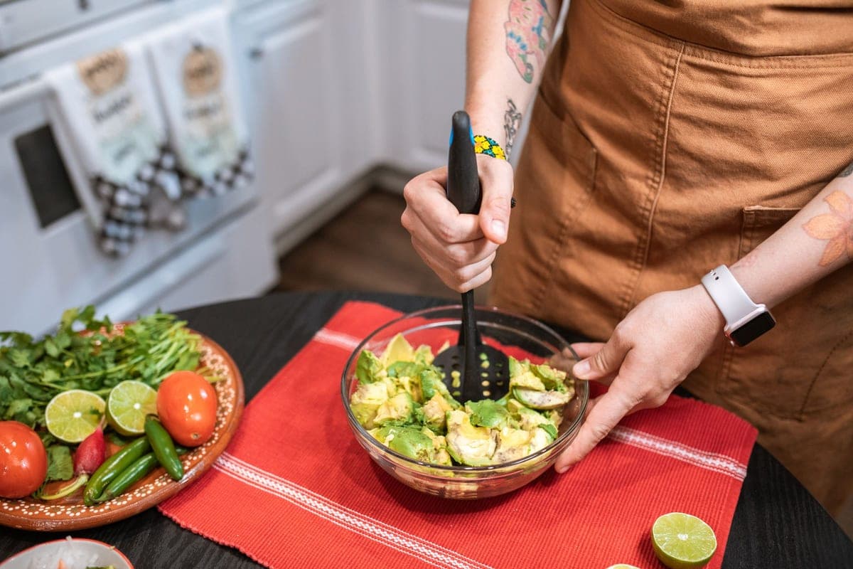 A cook in an apron mashing avocados in a glass bowl on a red cloth, with a plate of tomatoes, limes and peppers beside it