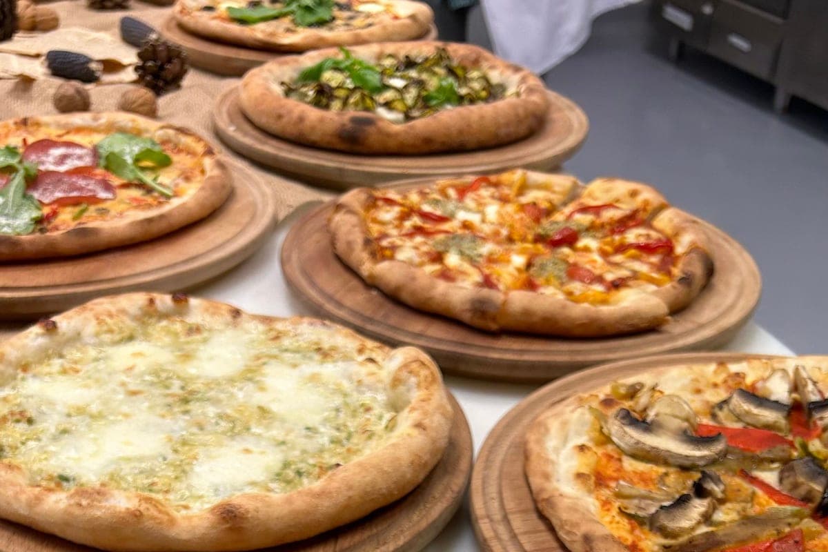 Several fresh homemade pizzas with puffy golden crusts on wooden boards, topped with tomato, mozzarella, mushrooms and greens