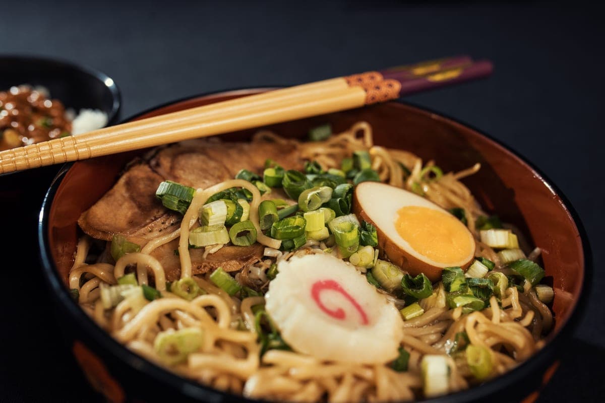A bowl of handmade shoyu ramen with wavy noodles, sliced pork, a halved marinated egg, fish cake and plenty of sliced scallions, with chopsticks resting on top