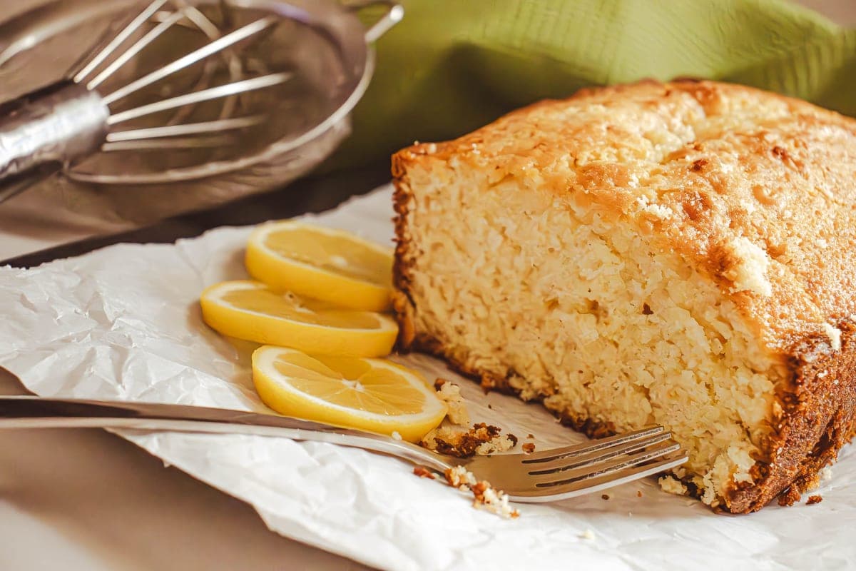 A golden lemon pound cake with a crackly top, cut to show its tender yellow crumb, beside fresh lemon slices and a fork
