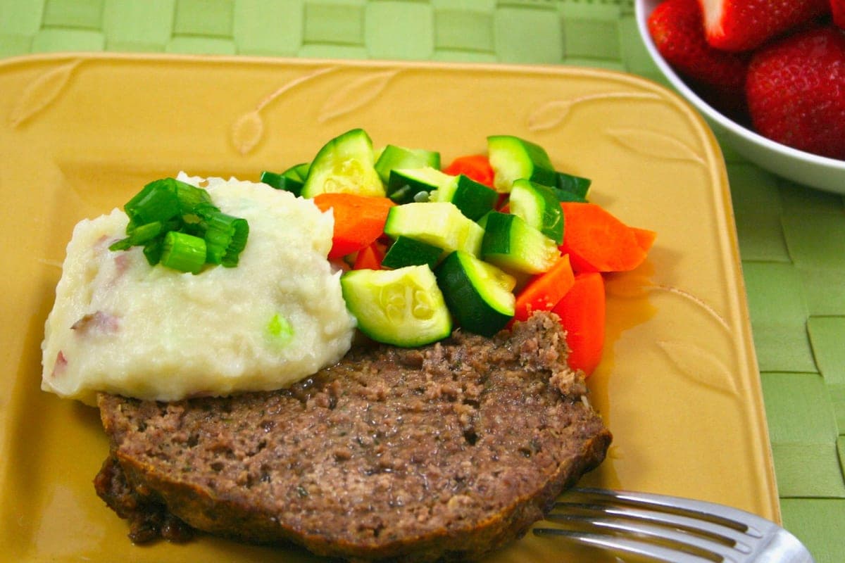 A slice of homemade meatloaf with mashed potatoes topped with green onions and a side of diced vegetables on a yellow plate