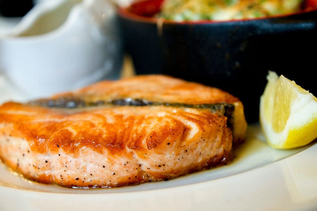 A golden pan-seared salmon fillet on a white plate with a lemon wedge, a small sauce pitcher and a salad behind