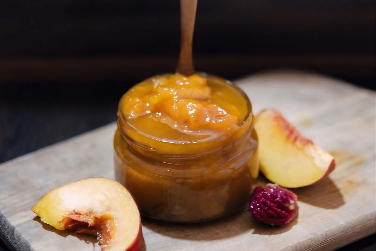 A small glass jar of golden peach jam with a wooden spoon, beside fresh peach slices on a wooden board