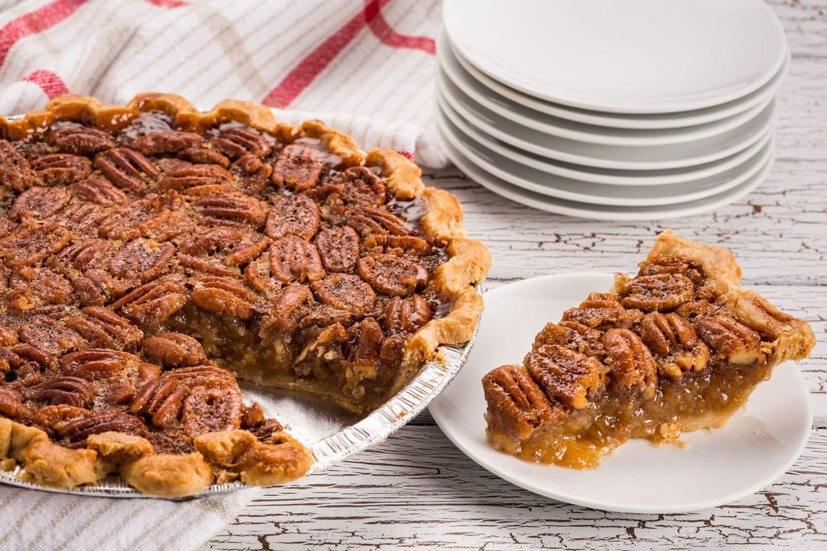 A pecan pie with one slice removed and served on a white plate beside a stack of plates