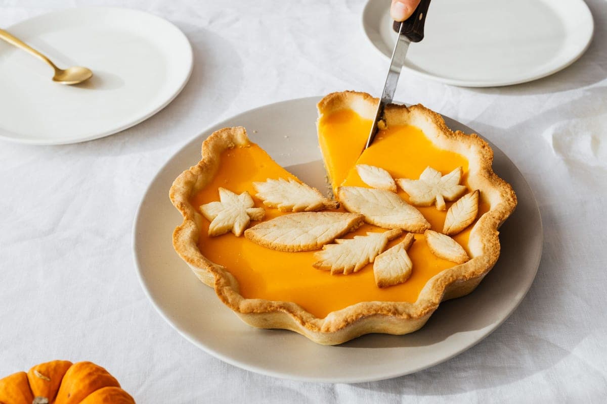 A pumpkin pie decorated with pastry leaves being sliced on a white tablecloth, with a small pumpkin and dessert plates nearby