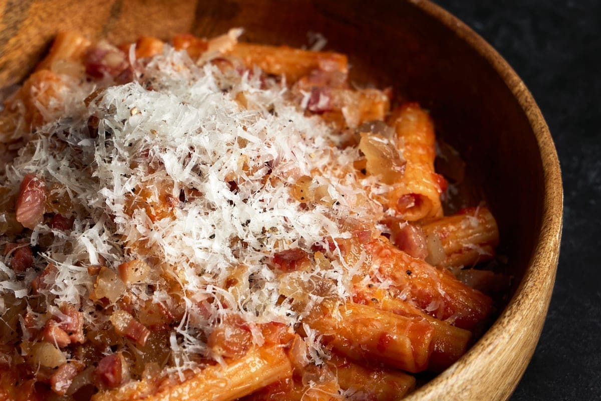 A wooden bowl of rigatoni in a rich tomato and pancetta sauce topped with a heap of finely grated cheese