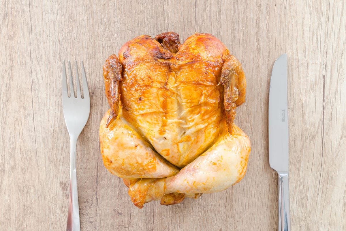 A whole golden-brown roast chicken seen from above on a pale wooden table, with a fork on one side and a knife on the other