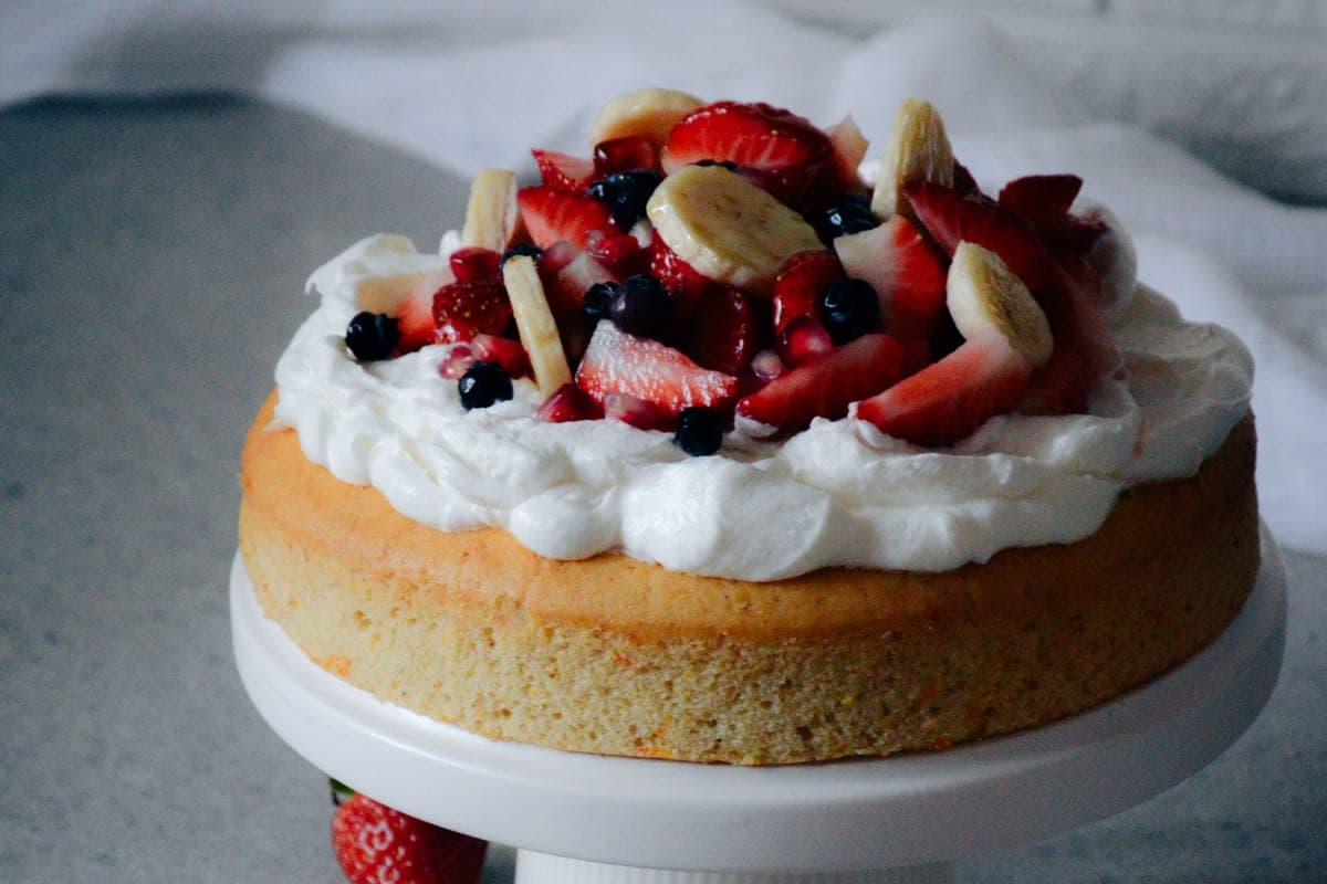 A golden vanilla cake on a white cake stand topped with whipped cream, sliced strawberries, blueberries and banana